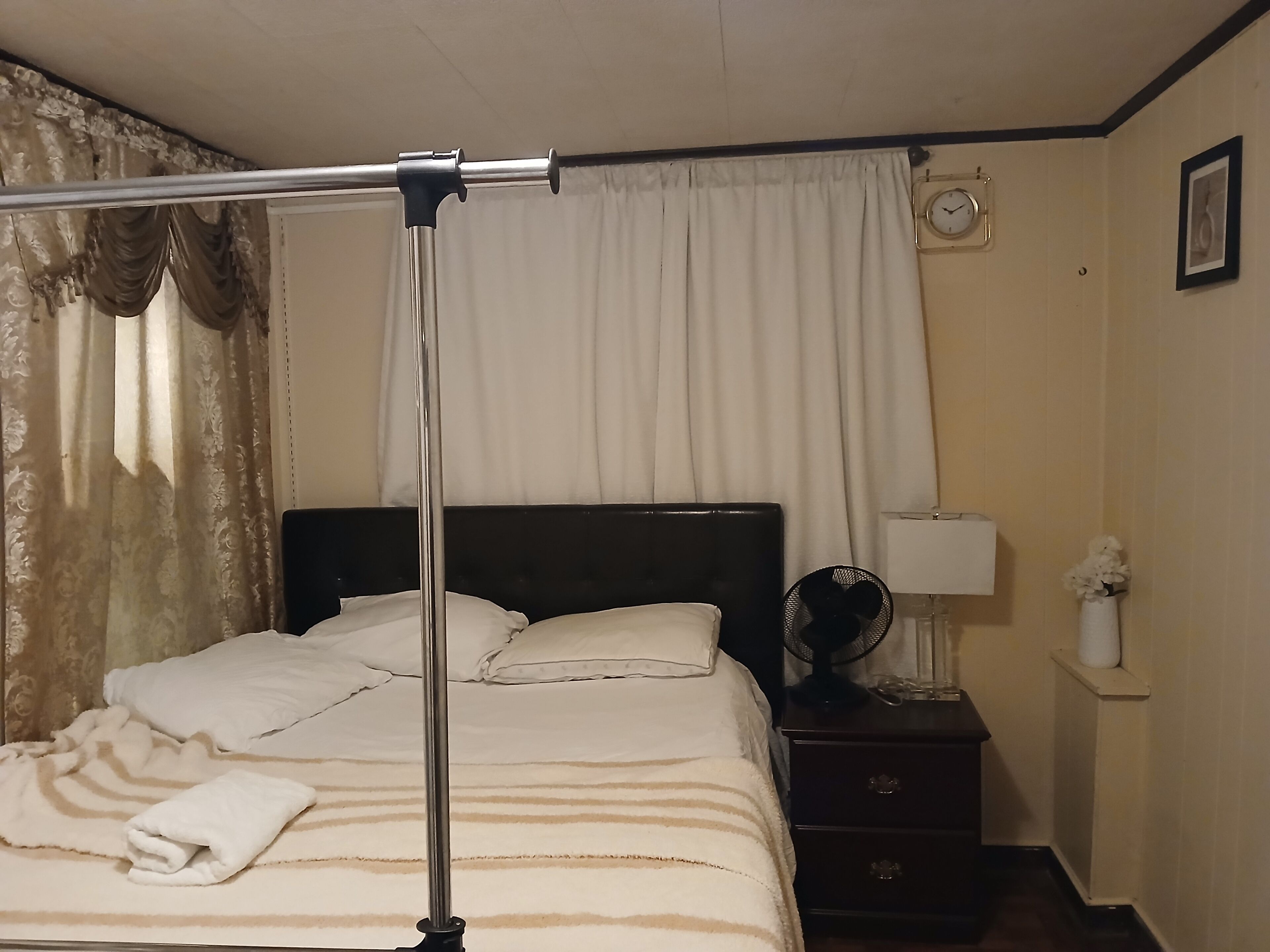 A neatly made bed with a striped blanket, a bedside table with a lamp, a clock on the wall, and a curtain partially drawn.