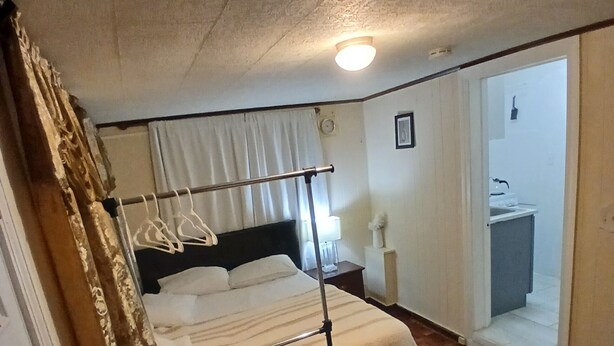 A bedroom with a bed, a nightstand, a hanging light fixture, a window with curtains, and a bathroom visible through an open door.