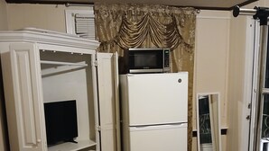 A compact room with a white refrigerator, a microwave on top, a television on a cabinet, and a wooden chair.