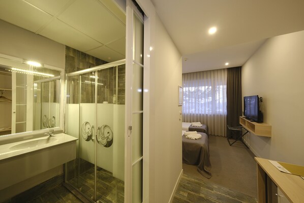 Habitación de hotel moderna con ducha de vidrio, un espejo grande, televisor de pantalla plana, mesita de noche y cama con cubrecama marrón.