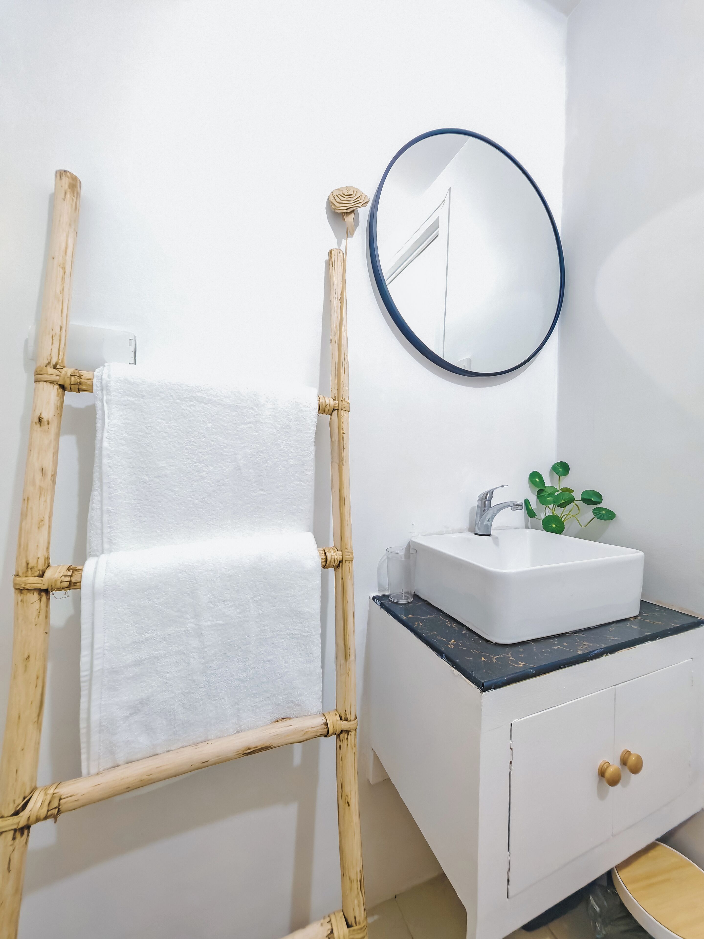 Un baño con tocador blanco, un espejo redondo, una toalla blanca en un perchero de bambú y una planta verde sobre el lavabo.
