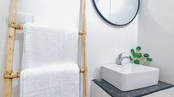 Un baño con tocador blanco, un espejo redondo, una toalla blanca en un perchero de bambú y una planta verde sobre el lavabo.