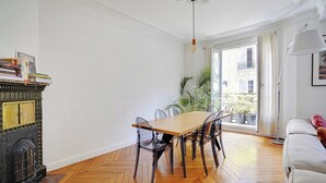 Une pièce dotée d’un parquet en bois disposé en motif à chevrons, d’une table à manger avec des chaises, d’une cheminée et d’une grande fenêtre offrant une vue sur les bâtiments environnants.