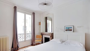 Une chambre d’hôtel avec un lit, une chaise, une cheminée, une grande fenêtre avec des rideaux et un luminaire au plafond.