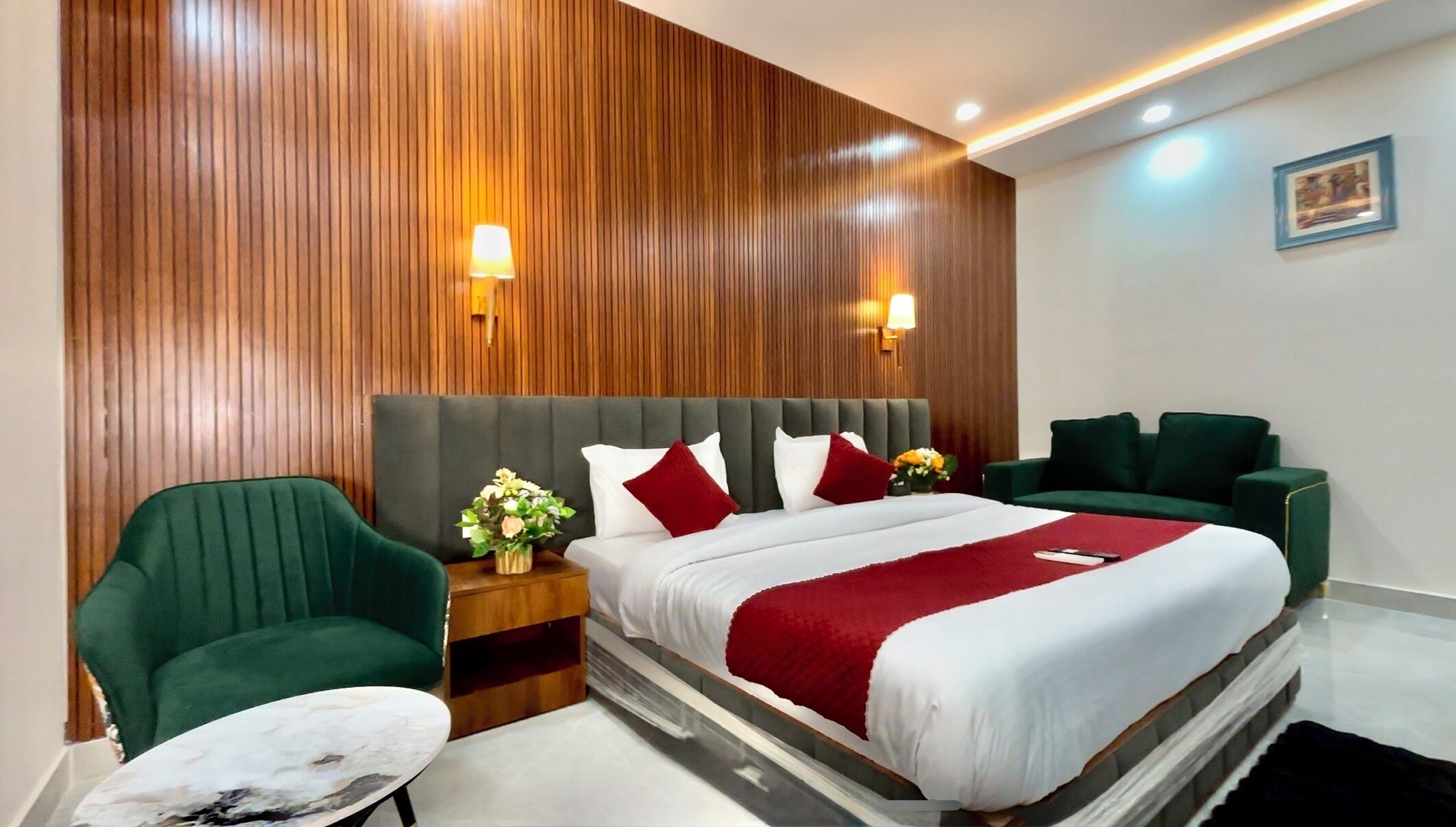 A hotel room with a large bed, two green armchairs, a small table, and a wooden wall panel.