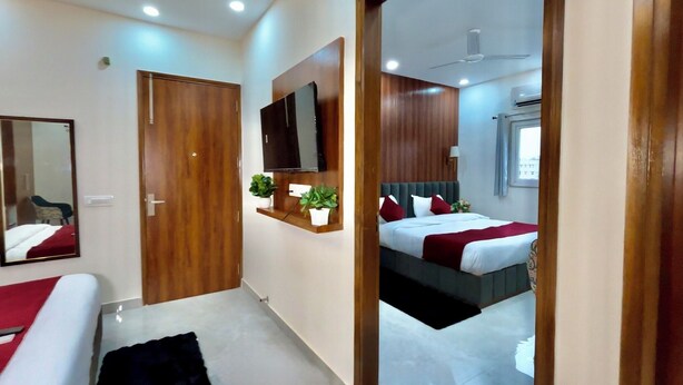 A hotel room with a wooden door, a flat-screen TV mounted on the wall, a bed with white bedding and red accents, a small potted plant, and a ceiling fan. - Hotel The Tusk (Rishikesh)