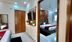 A hotel room with a wooden door, a flat-screen TV mounted on the wall, a bed with white bedding and red accents, a small potted plant, and a ceiling fan. - Hotel The Tusk (Rishikesh)