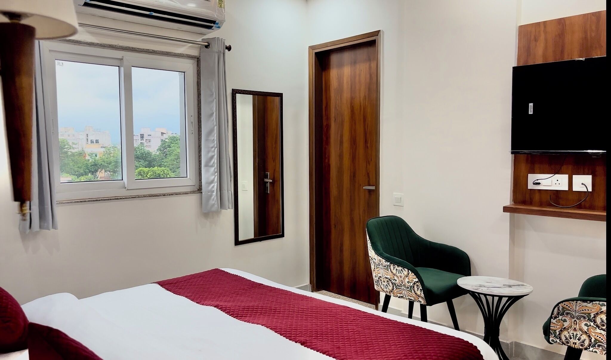 A hotel room with a bed, a chair, a small table, a TV, and a window with a view of buildings.