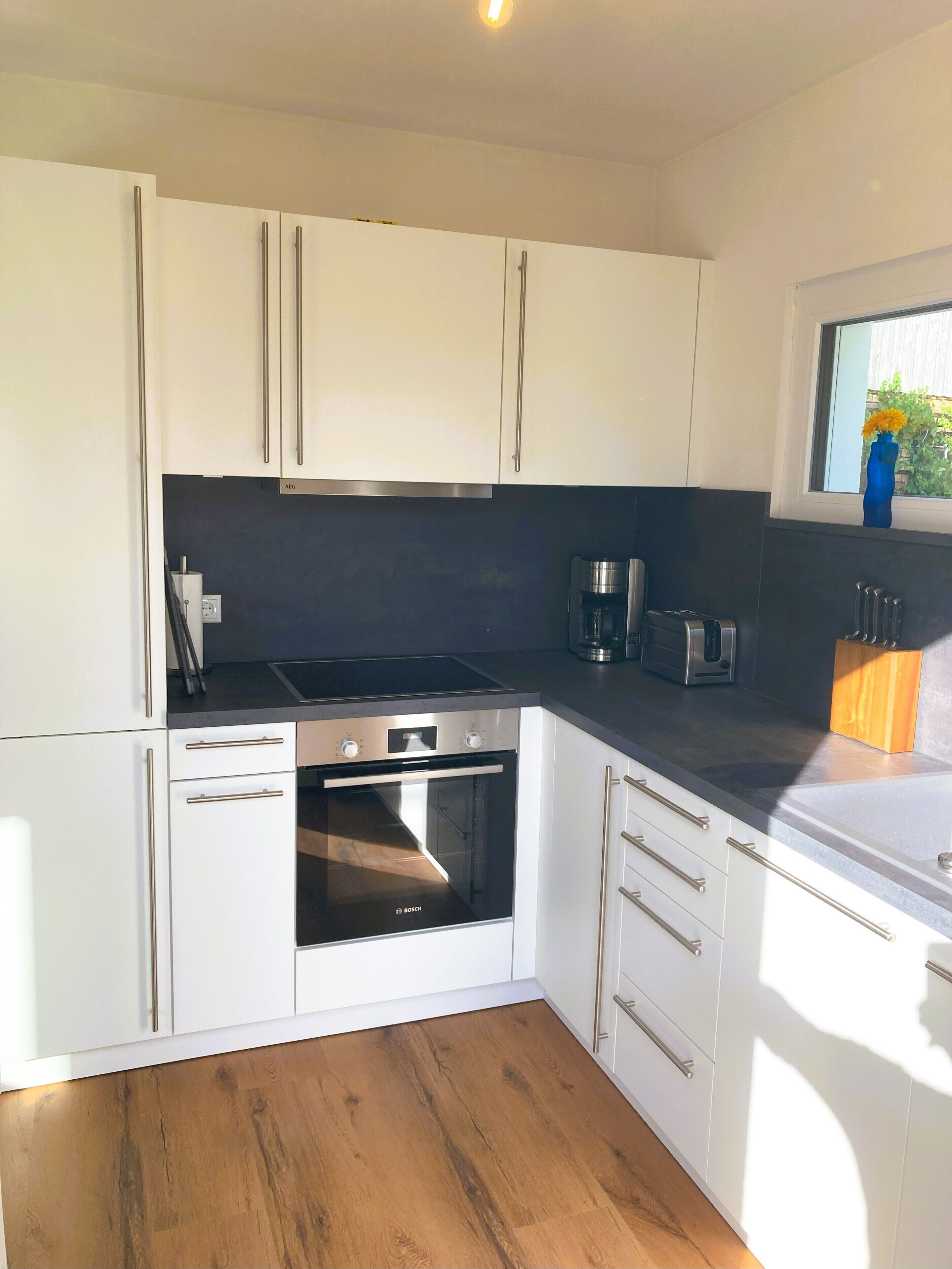 Una cocina moderna con armarios blancos, un horno Bosch, una cafetera y un ventanal con vistas al verdor.
