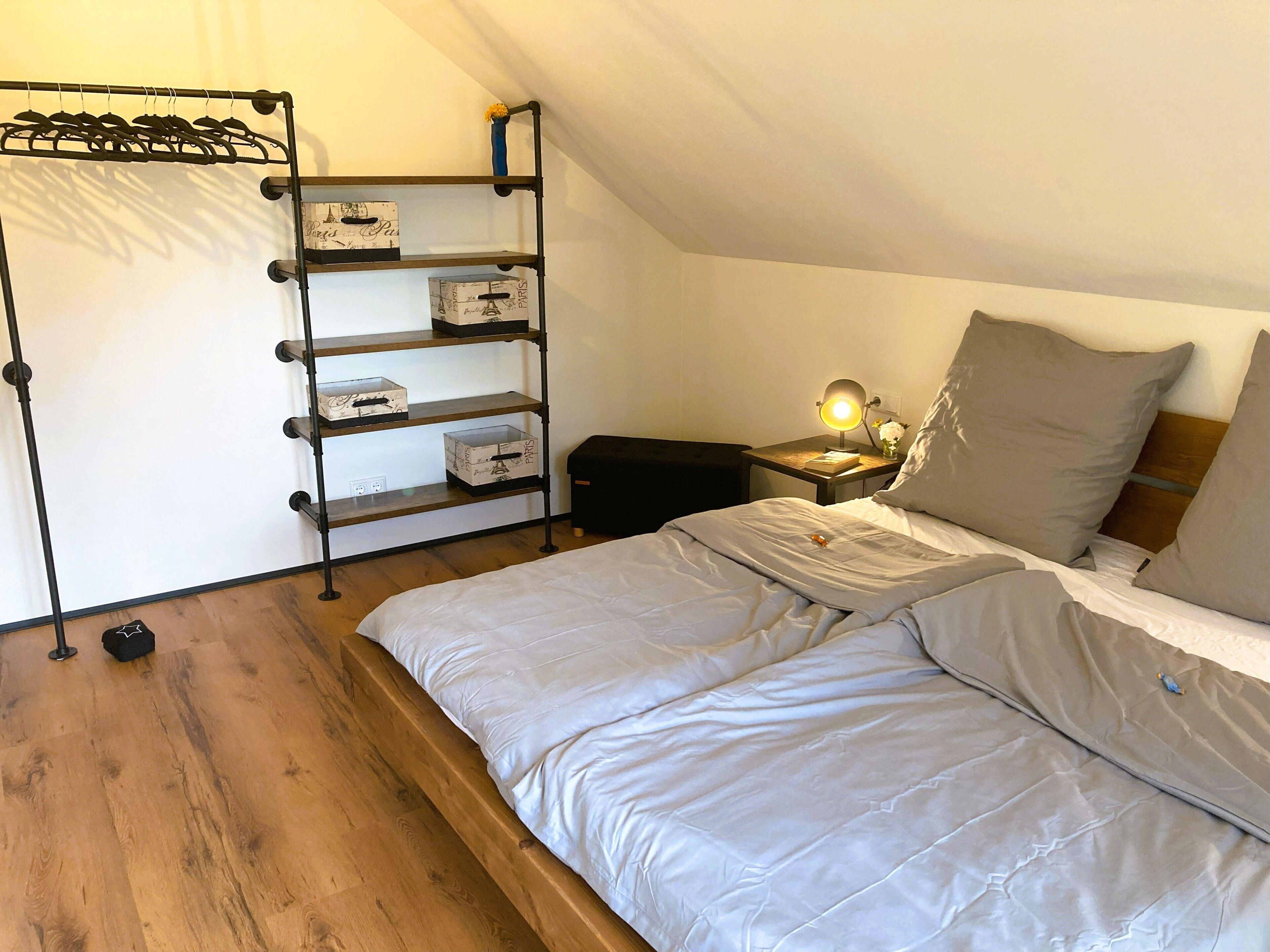 Une chambre à coucher comprenant un lit, une table de chevet avec une lampe, une étagère avec des boîtes de rangement et un porte-manteau.