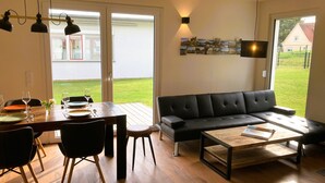 A modern living room with a wooden dining table, black chairs, a black sofa, and a coffee table. There is a sliding glass door leading to a garden. - Riedlodge Lake Constance-Upper Swabia Holiday Home (Guggenhausen)