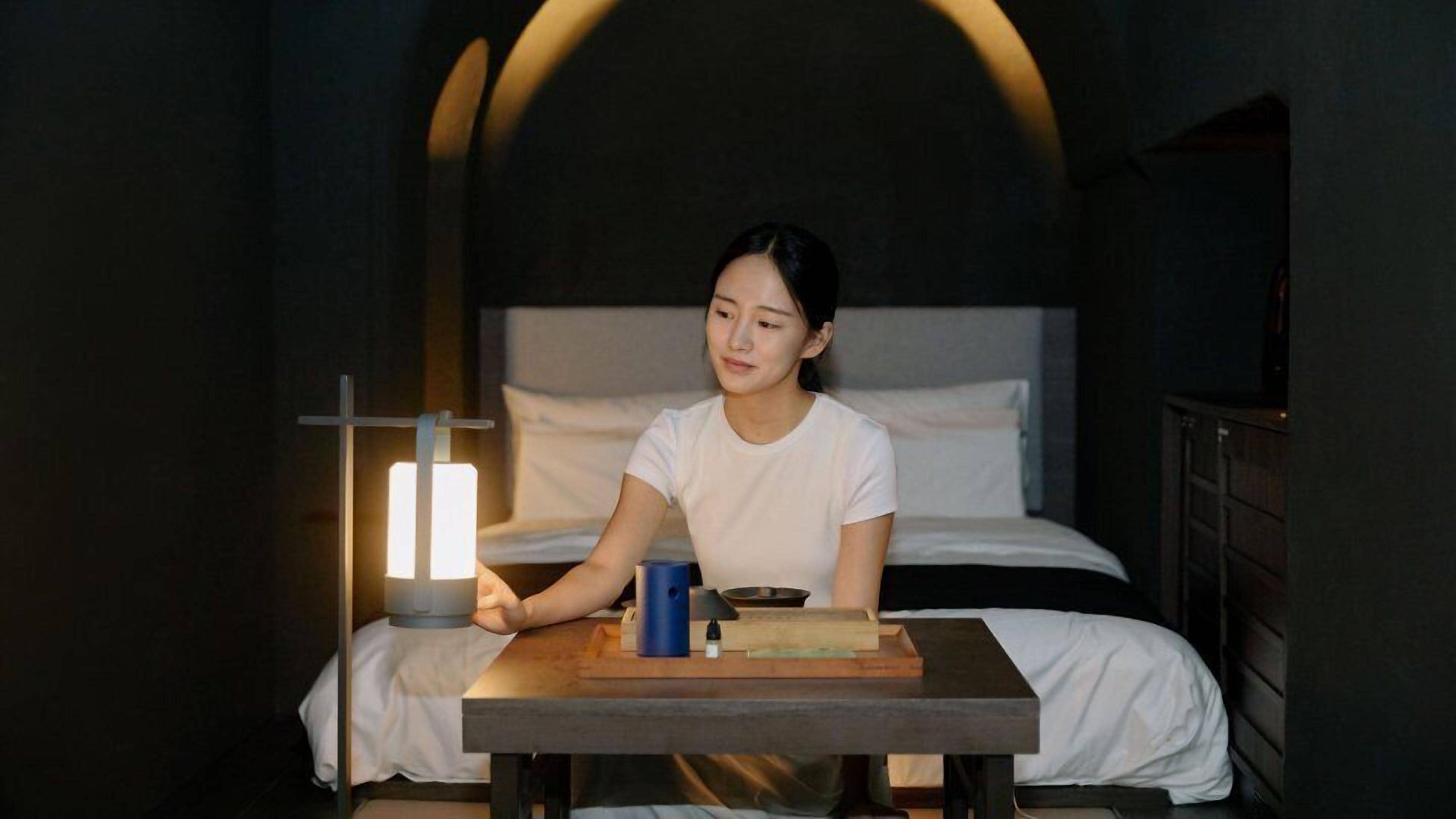Una persona sentada a una mesa en una habitación con poca luz, con una cama al fondo.