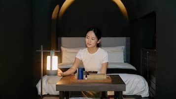 Una persona sentada a una mesa en una habitación con poca luz, con una cama al fondo.