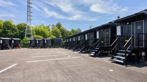 A row of black portable cabins with numbered doors, arranged in a parking lot with a clear sky and trees in the background. - HOTEL R9 The Yard Mizunami (Mizunami)