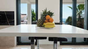 Un espace repas moderne avec une table blanche, un panier de fruits contenant des ananas, des bananes et des oranges, et de grandes fenêtres offrant une vue sur les palmiers et la mer.