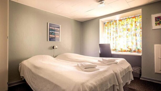 Une chambre d’hôtel avec deux lits, une fenêtre avec des rideaux colorés, une chaise et une affiche accrochée au mur.