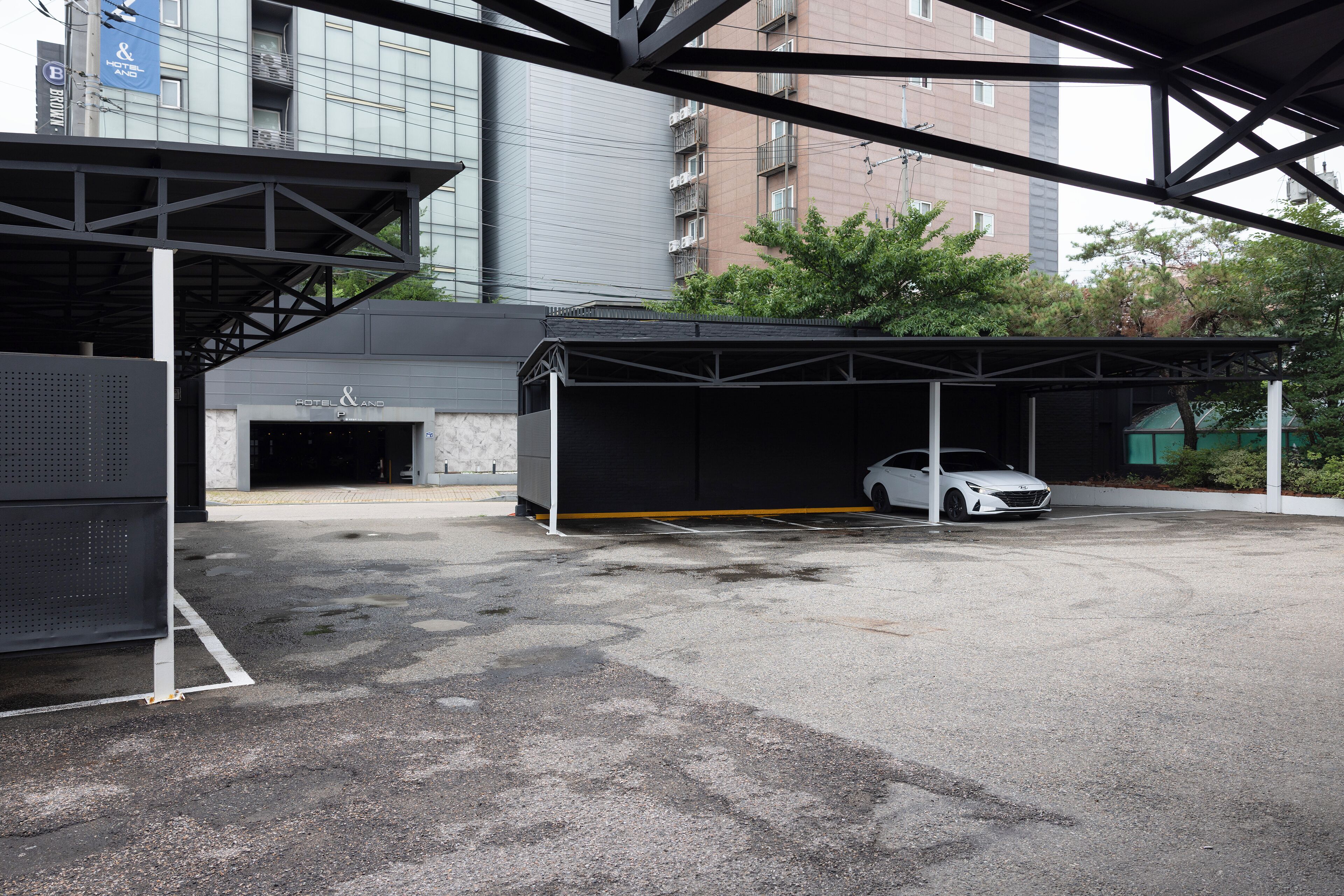 A covered parking area with a white car inside, a building with a sign reading "HOTEL & PARK", and a modern building in the background.