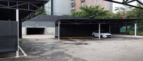 A covered parking area with a white car inside, a building with a sign reading "HOTEL & PARK", and a modern building in the background.