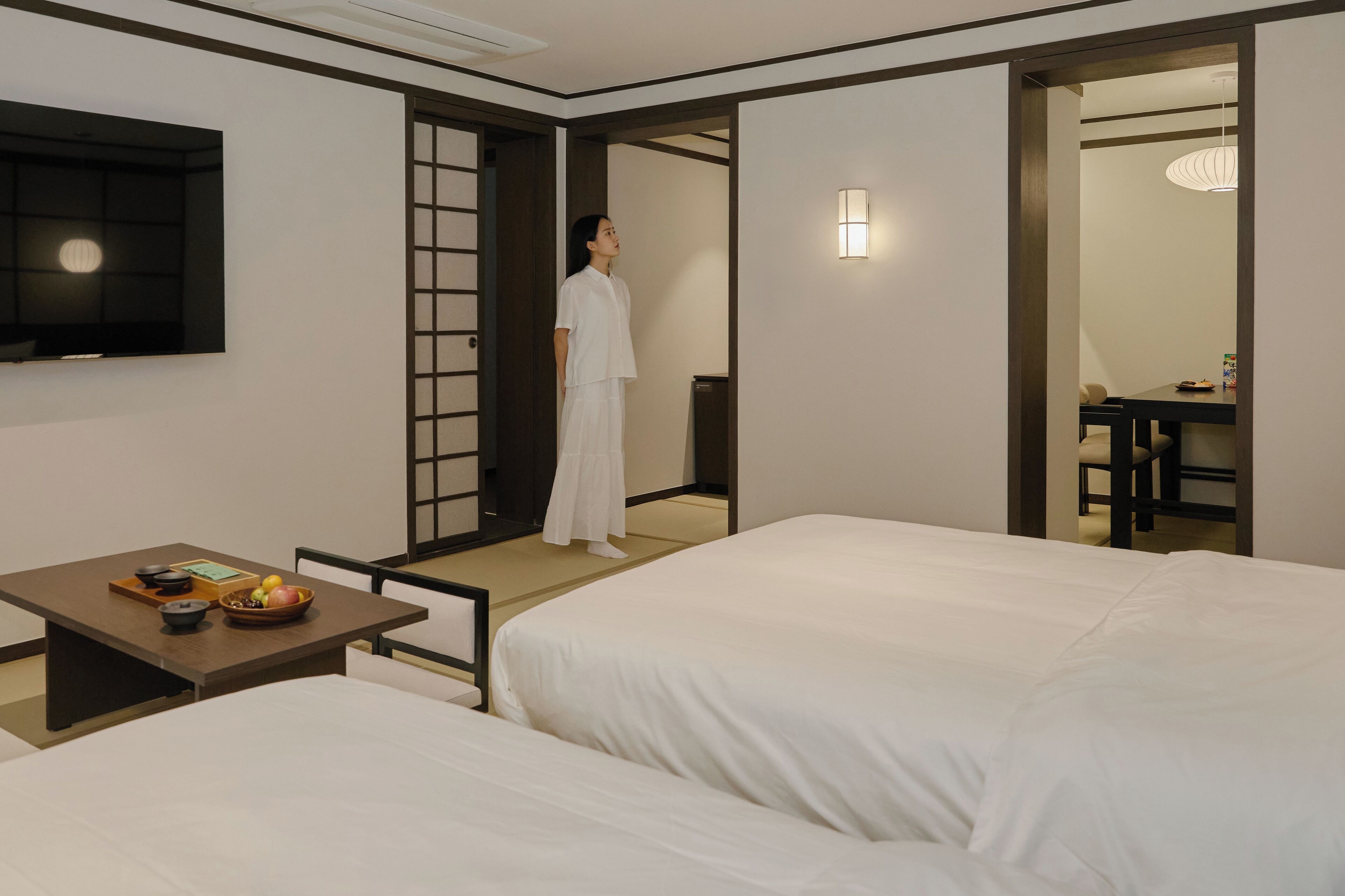 A hotel room with two beds, a dining table with a bowl of fruit, and a TV on the wall.