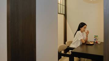 A person seated at a table with a beverage and a box, in a room with a wooden floor and a ceiling light fixture.