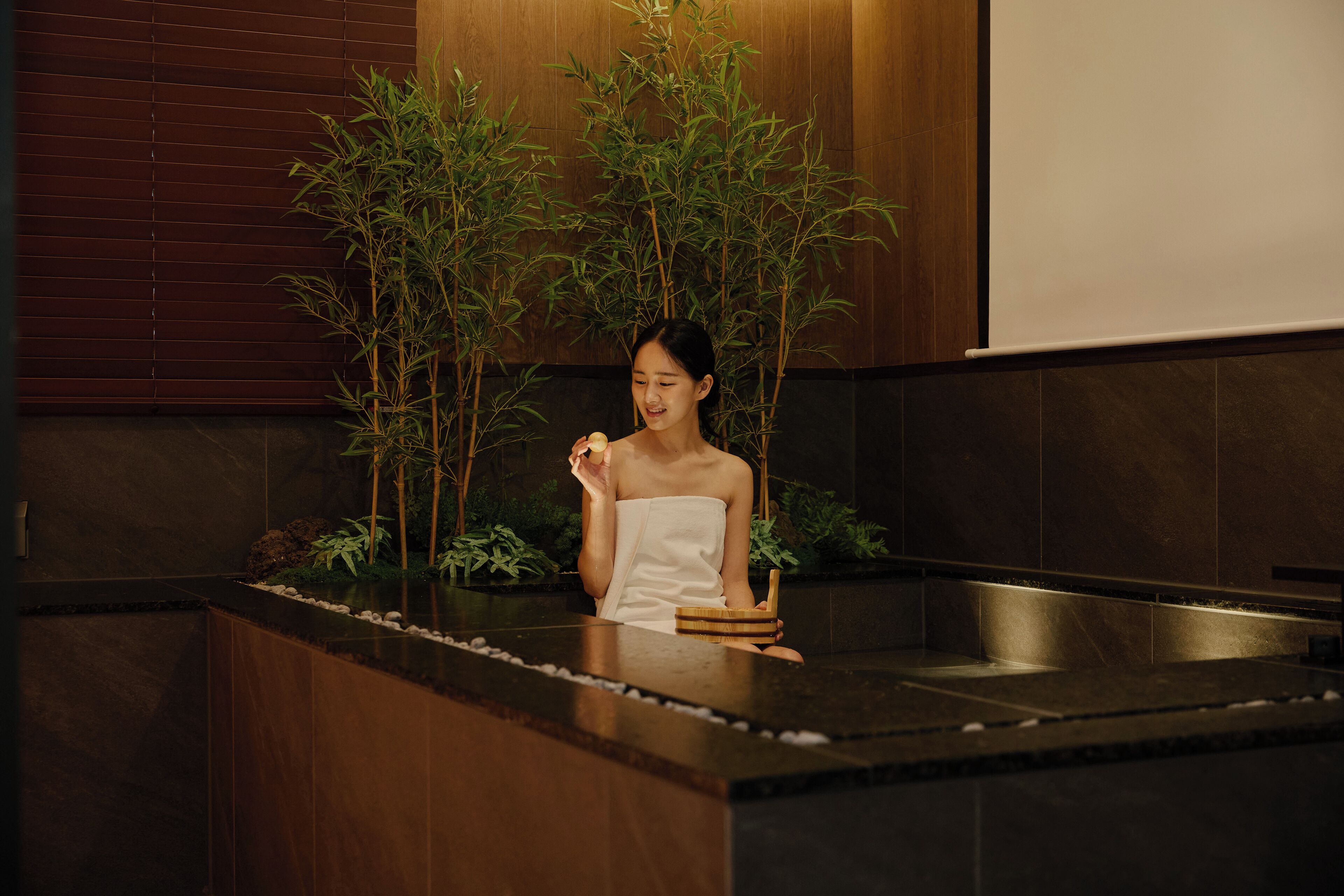 Un espacio de spa con un mostrador de recepción de madera, plantas de bambú y una persona sentada en una bañera.