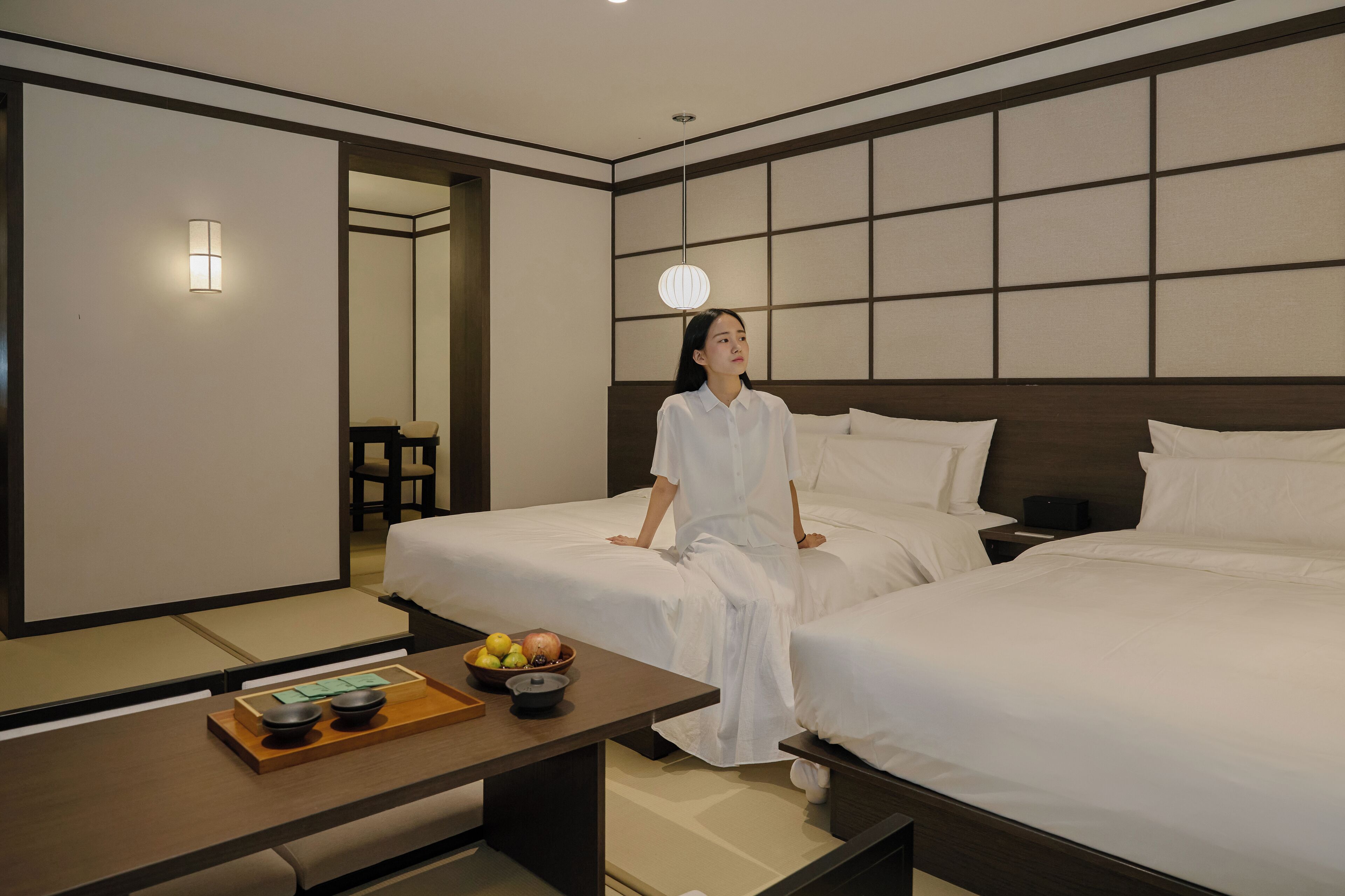 A hotel room with two beds, a wooden table with a fruit bowl, and a wall-mounted light fixture.