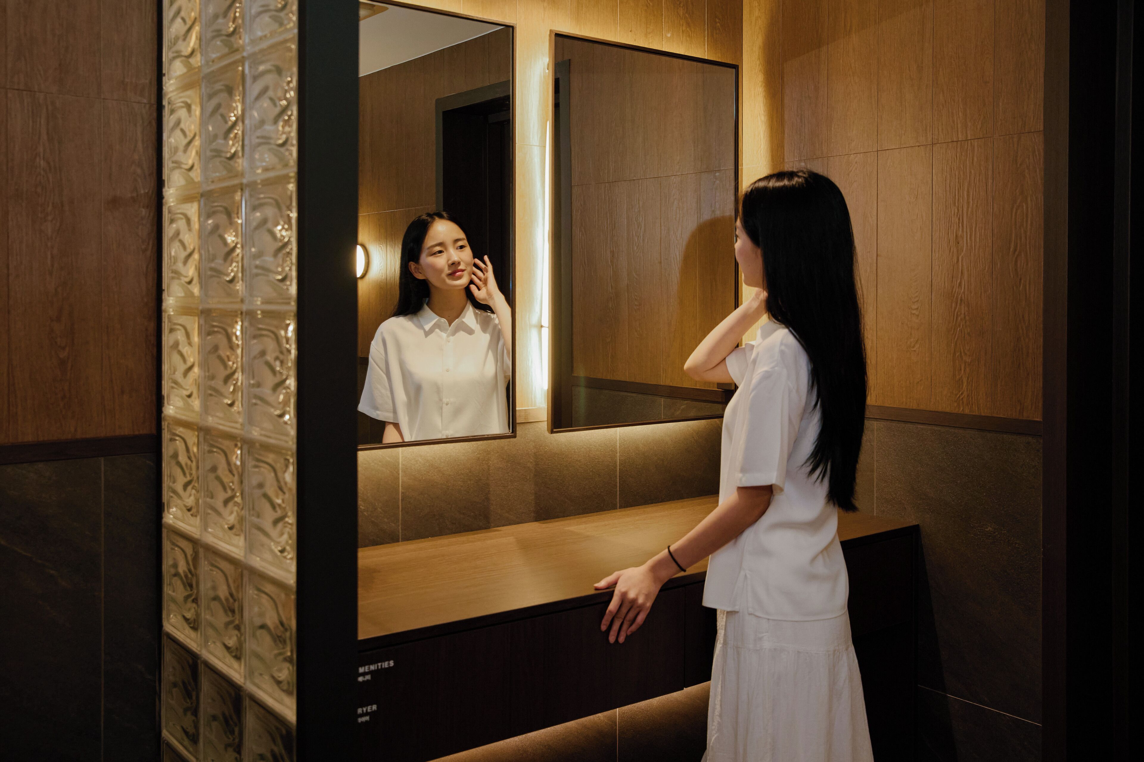 A woman in a white dress standing in front of a mirror in a room with wooden paneling and a tiled floor.