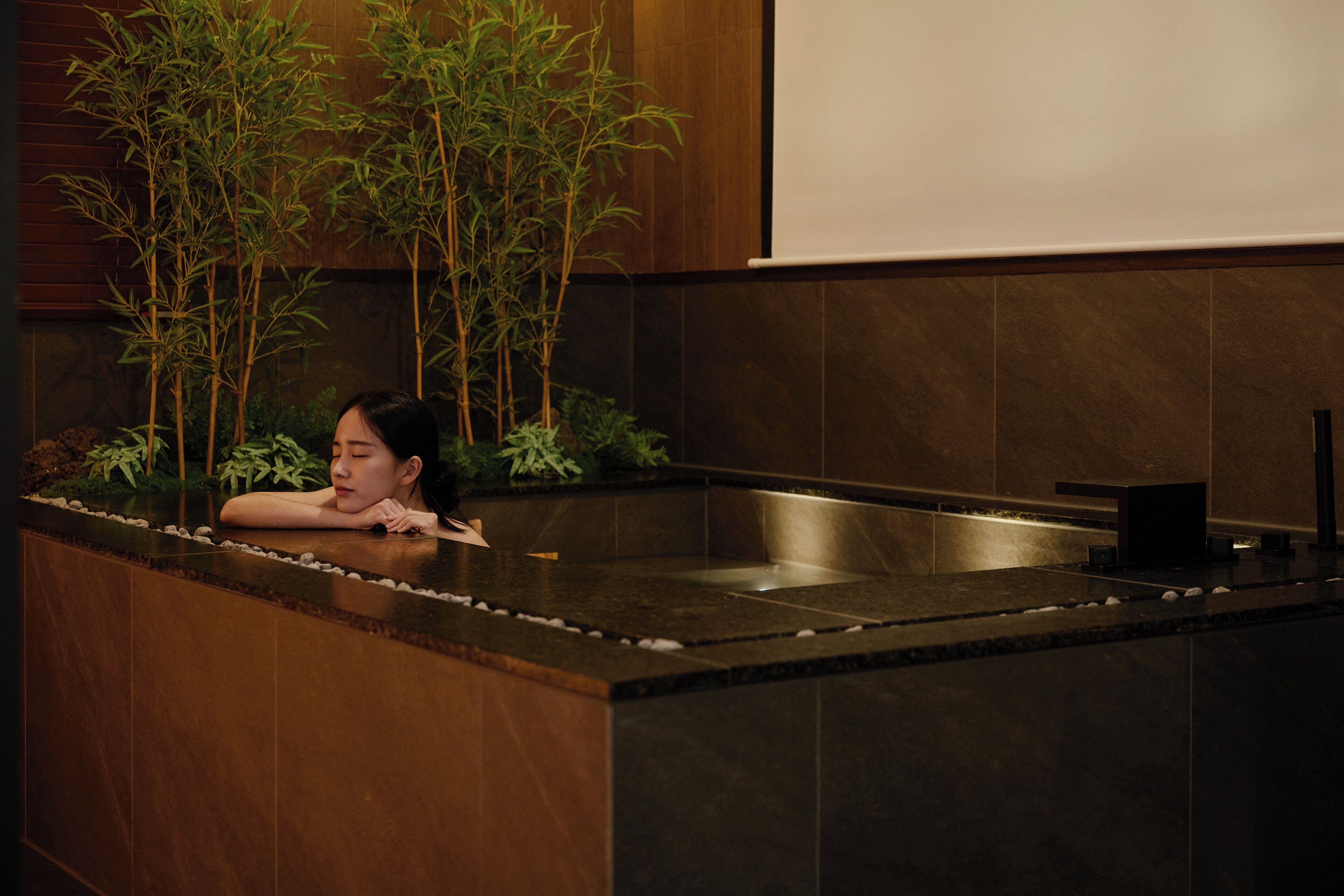 Una mujer descansando sobre una encimera con un lavabo y plantas al fondo.