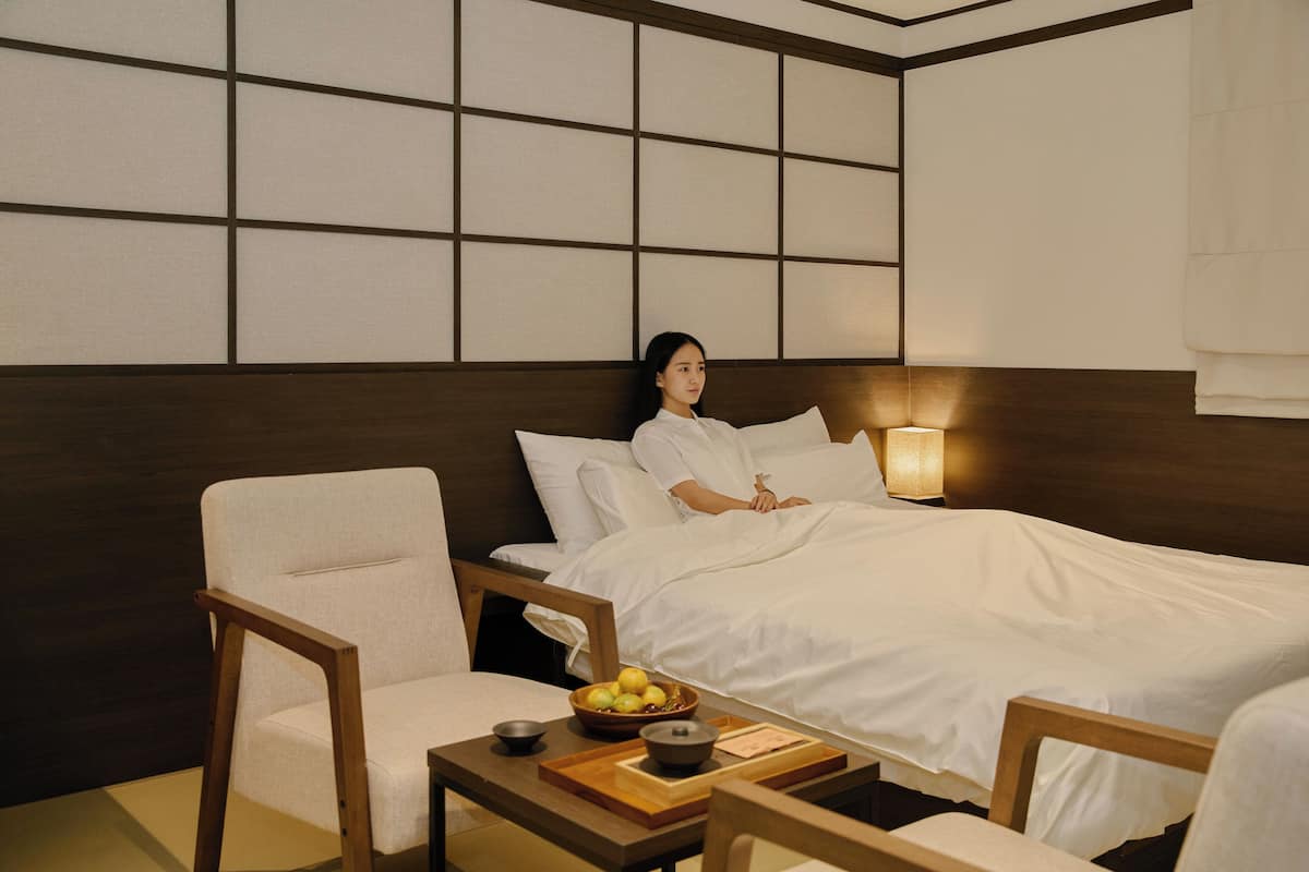 A hotel room with a bed, a bedside table with a bowl of fruit, a chair, and a wall with a grid pattern.