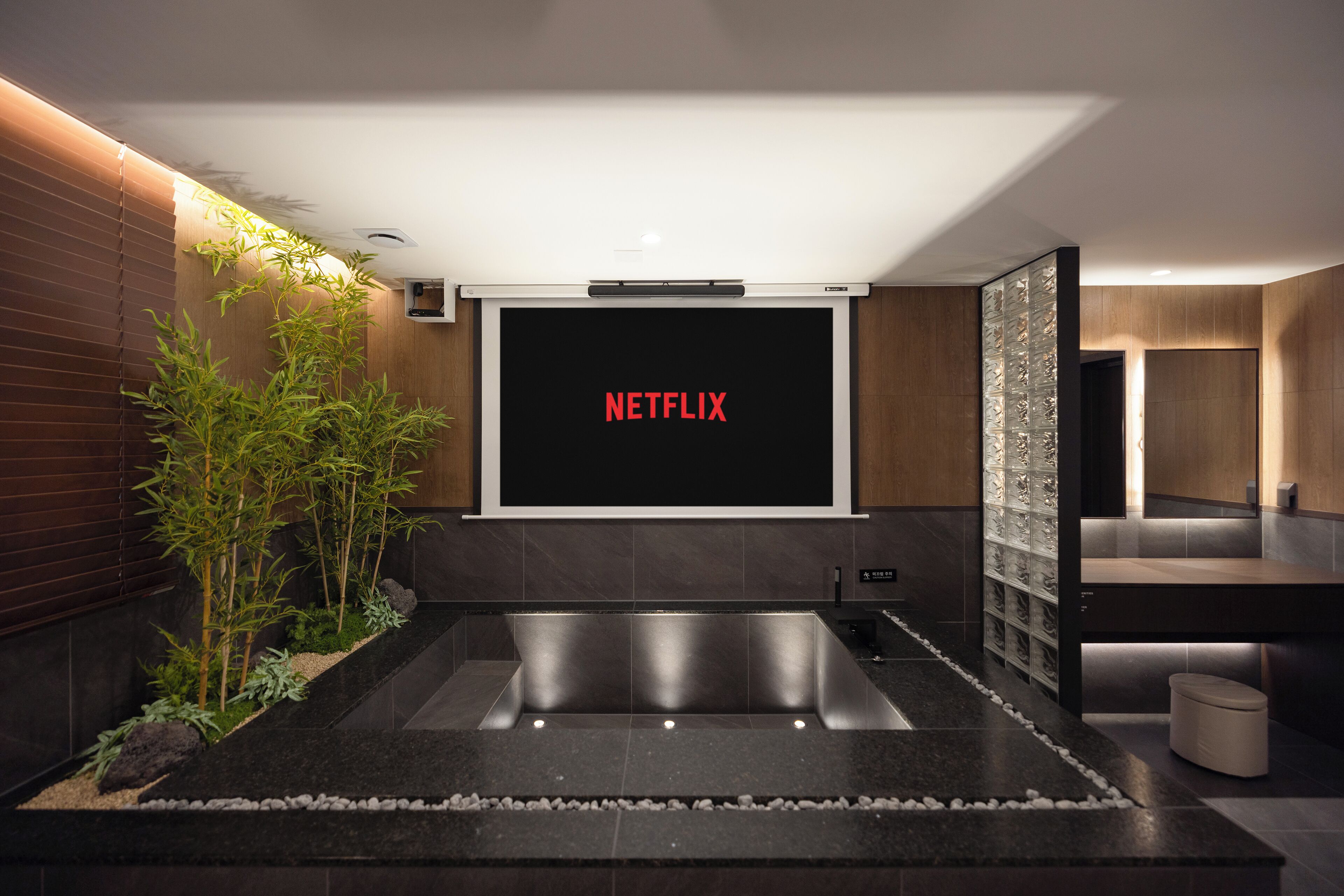Ein modernes Badezimmer mit einem gro ßen Spiegel, einem Waschbecken und einer Toilette. Ein an der Wand montierter Fernseher zeigt das Netflix-Logo.