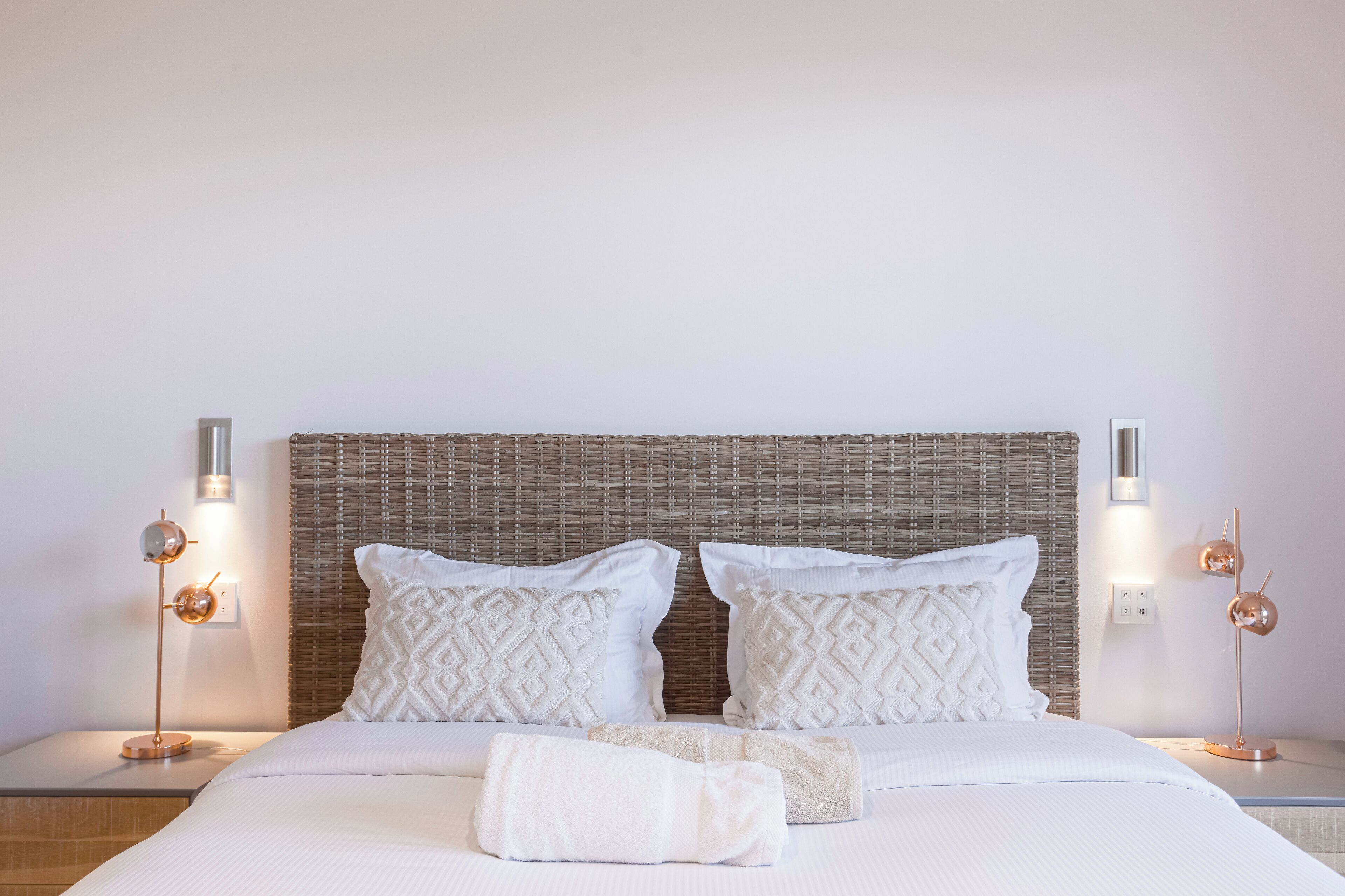 Un lit bien fait, avec du linge de lit blanc et deux coussins décoratifs, flanqué de deux tables de chevet surmontées de lampes modernes.