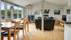 Een ruime woonkamer met een eettafel, stoelen, een bank en een televisie. Grote ramen zorgen voor natuurlijk licht en uitzicht op de omgeving.