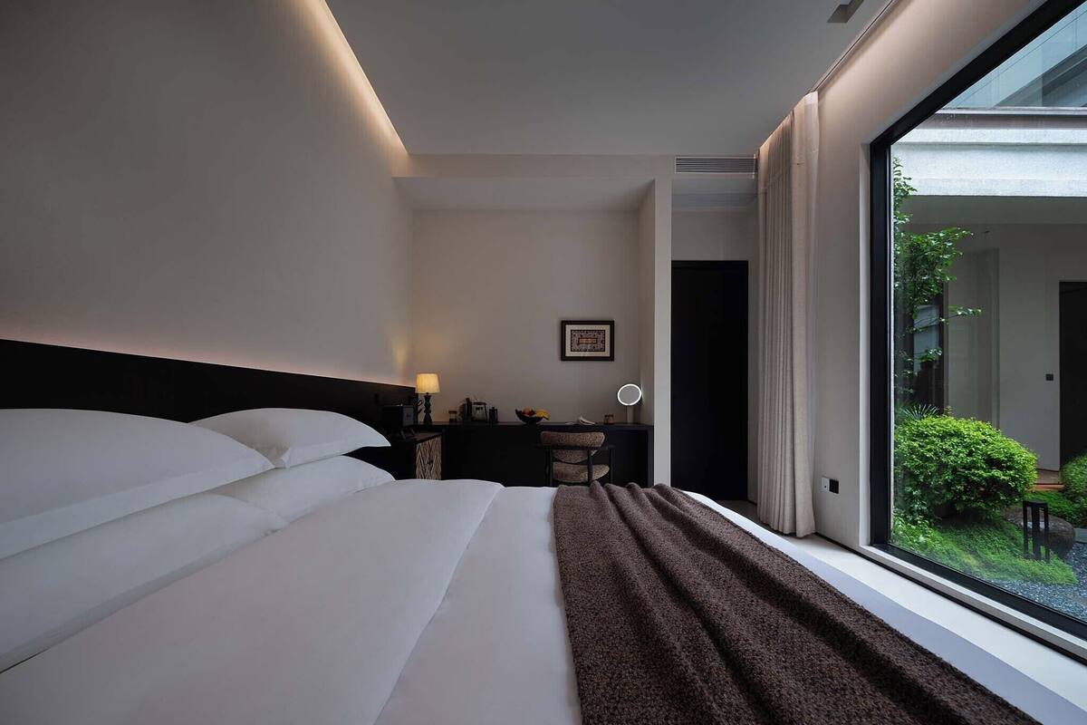 Une chambre d’hôtel avec un grand lit, un bureau avec une chaise, une petite table avec une lampe et une vue sur la verdure par la fenêtre.