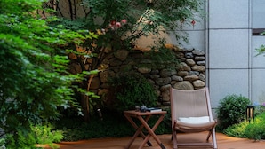 A wooden deck with a wicker chair and a small table, surrounded by lush greenery and a stone wall. - Qi Yi Nanxu Pavilion Homestay (Kunming)