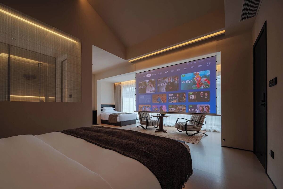 Une chambre d’hôtel moderne dotée d’un grand lit, d’un coin salon avec deux fauteuils, d’un écran de télévision diffusant du contenu et d’une grande fenêtre.