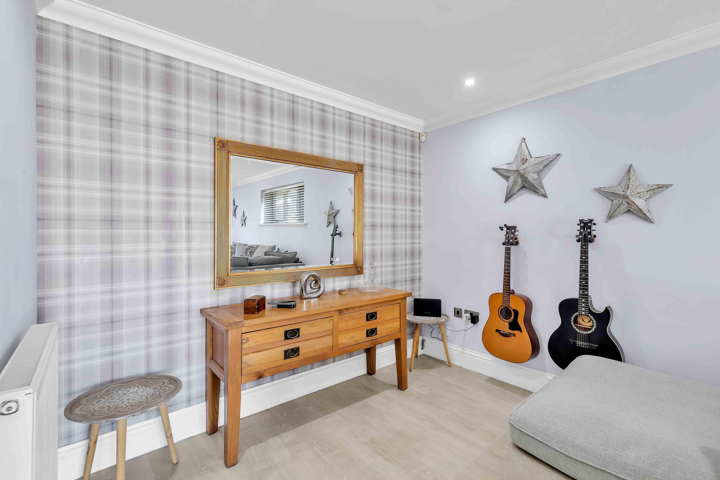 A bedroom with a plaid wallpaper, a wooden dresser, a mirror, two guitars, and a small stool.