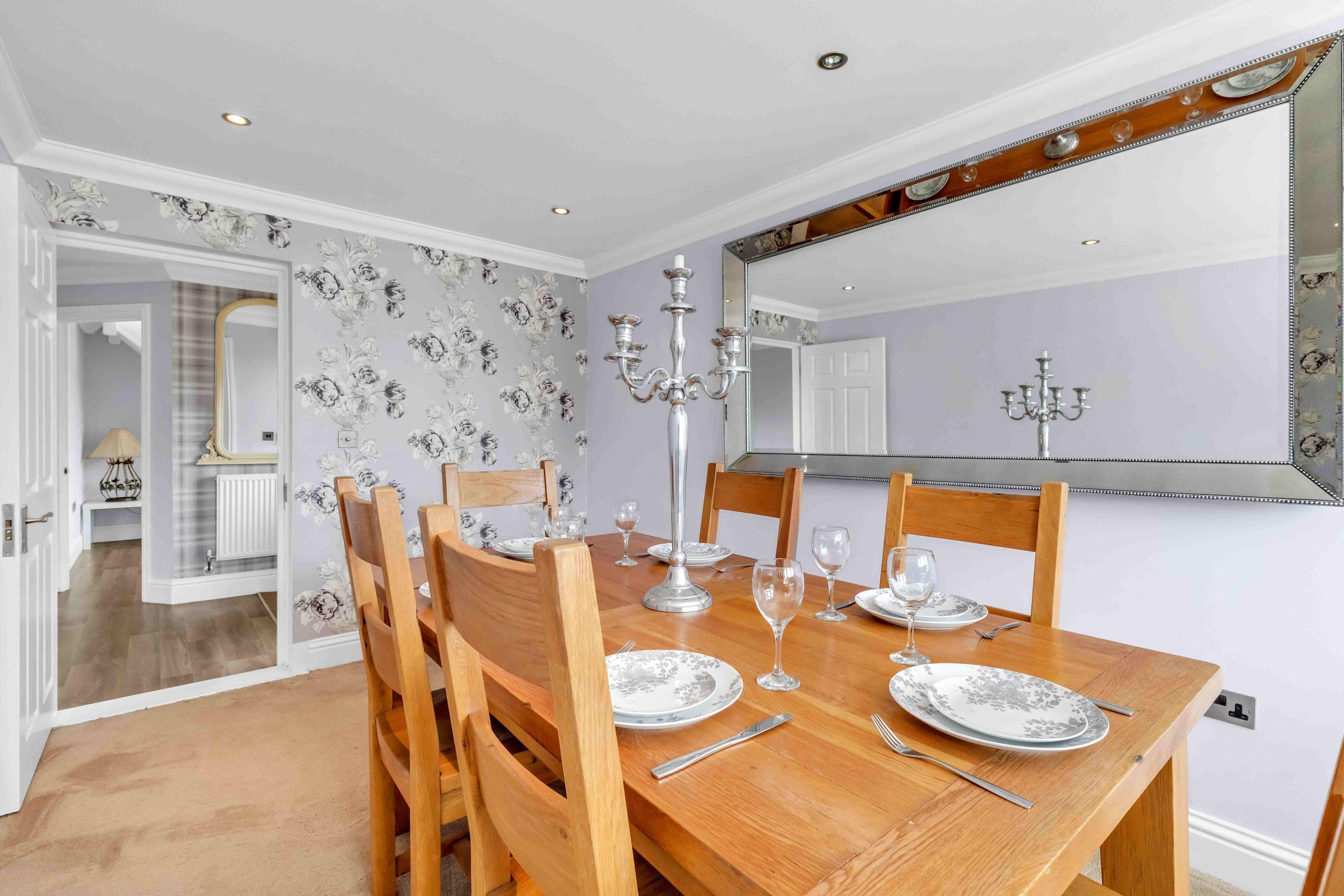 Une salle à manger avec une table en bois dressée pour quatre personnes, entourée de chaises. Un lustre est suspendu au-dessus de la table, et un grand miroir reflète la pièce.