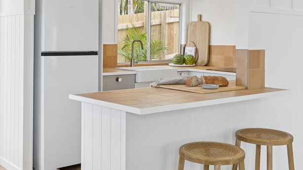 A kitchen with a white countertop, wooden stools, and a window with a view of a wooden fence and greenery.