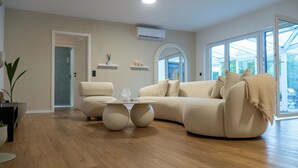 A modern living room with a beige sofa, a round coffee table, and a large window providing a view of the outdoors. - VILLAFINE - Exclusive villa with pool & sauna in Vaihingen an der Enz (Aurich, Vaihingen an der Enz)