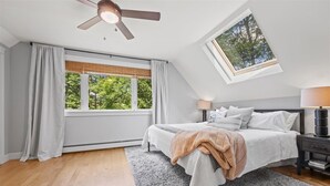 Une chambre à coucher comprenant un lit, des tables de chevet, un ventilateur de plafond, une fenêtre donnant sur des arbres et un puits de lumière.
