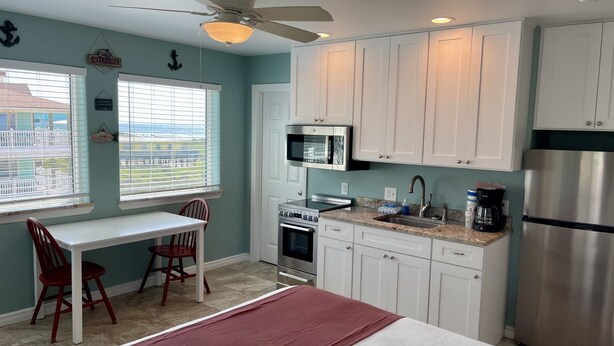 A compact kitchen with white cabinets, a microwave, stove, and refrigerator. There is a dining area with a table and chairs, and a window with blinds. - Beachgate Condo Suites And Hotel 232 1 Bedroom Condo (Port Aransas)