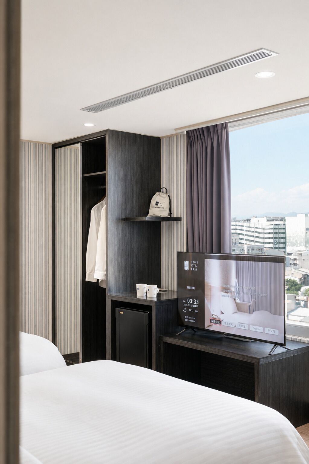 A hotel room with a bed, a TV, a small desk, a wardrobe, and a window with curtains.