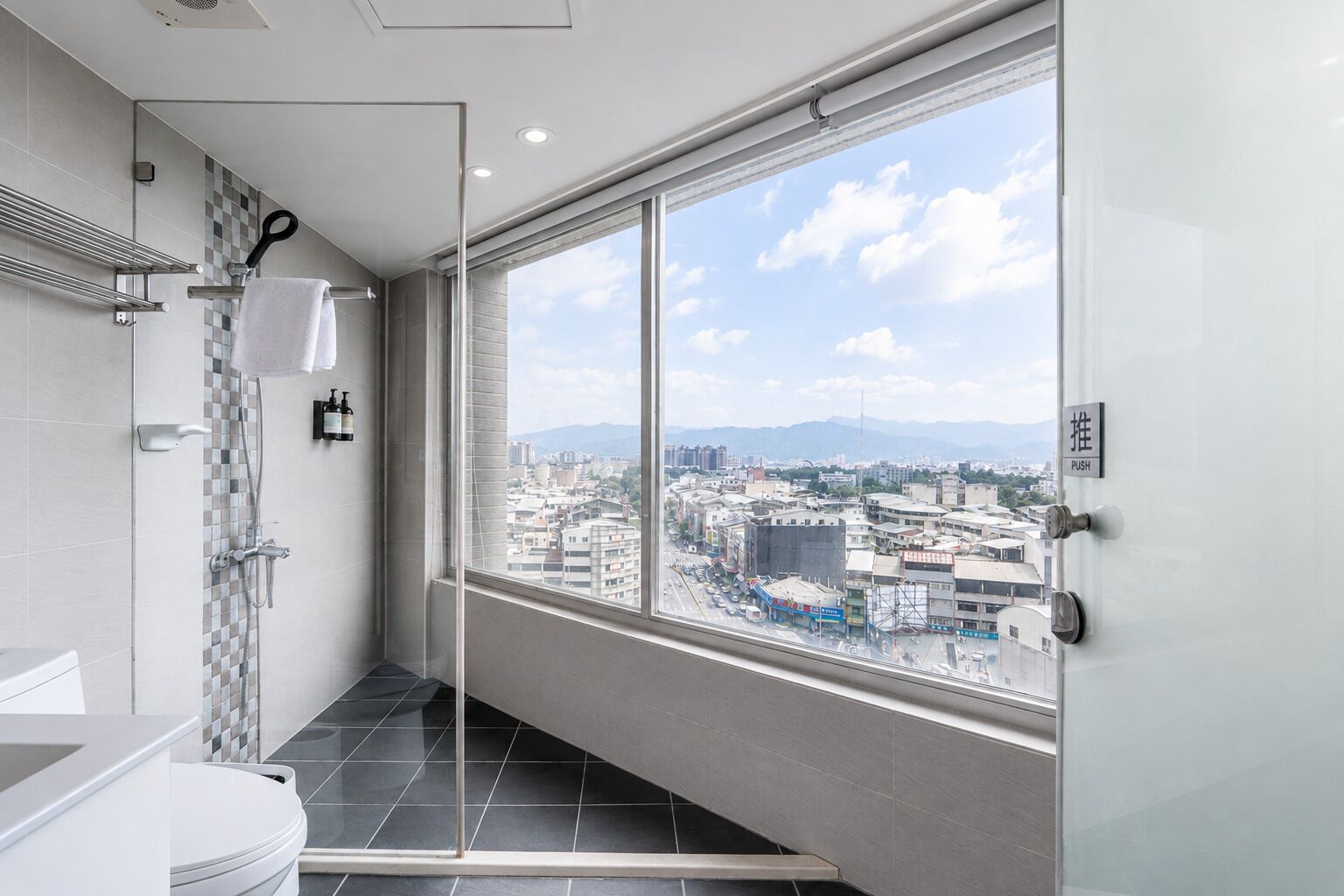 A modern bathroom with a shower, toilet, and a large window offering a city view.