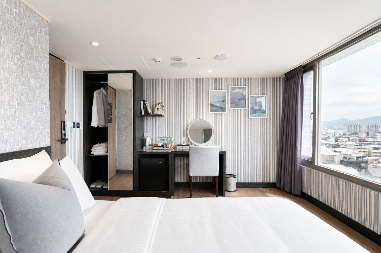 A hotel room with a bed, a desk with a mirror, a wardrobe, and a city view through a large window.