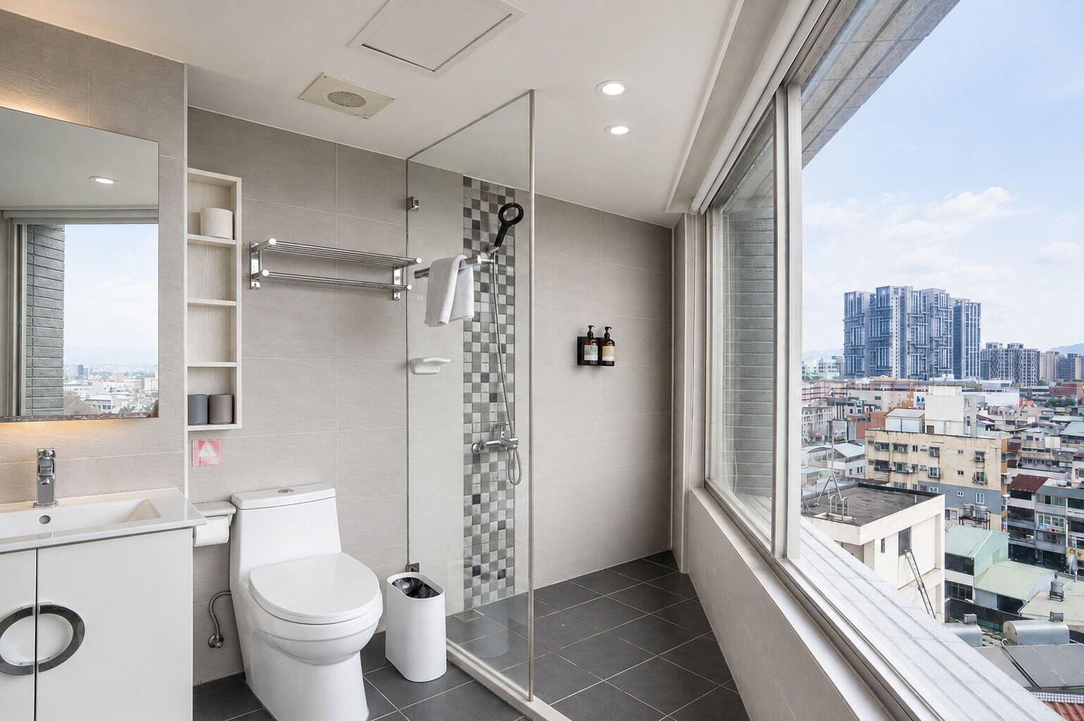 A modern bathroom with a toilet, sink, and shower. It has a large window with a city view.