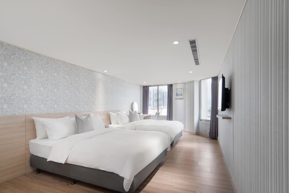 A modern hotel room with a large bed, a flat-screen TV mounted on the wall, and a city view through large windows.