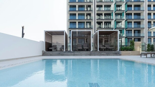 A swimming pool with lounge chairs and a modern building with balconies in the background. - Bloomfields Cozy 1Br Binghatti House  (Dubai)