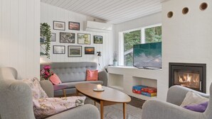 A modern living room with a grey sofa, two armchairs, a coffee table, and a fireplace. There are framed pictures on the wall, a TV, and a window with a view of trees.