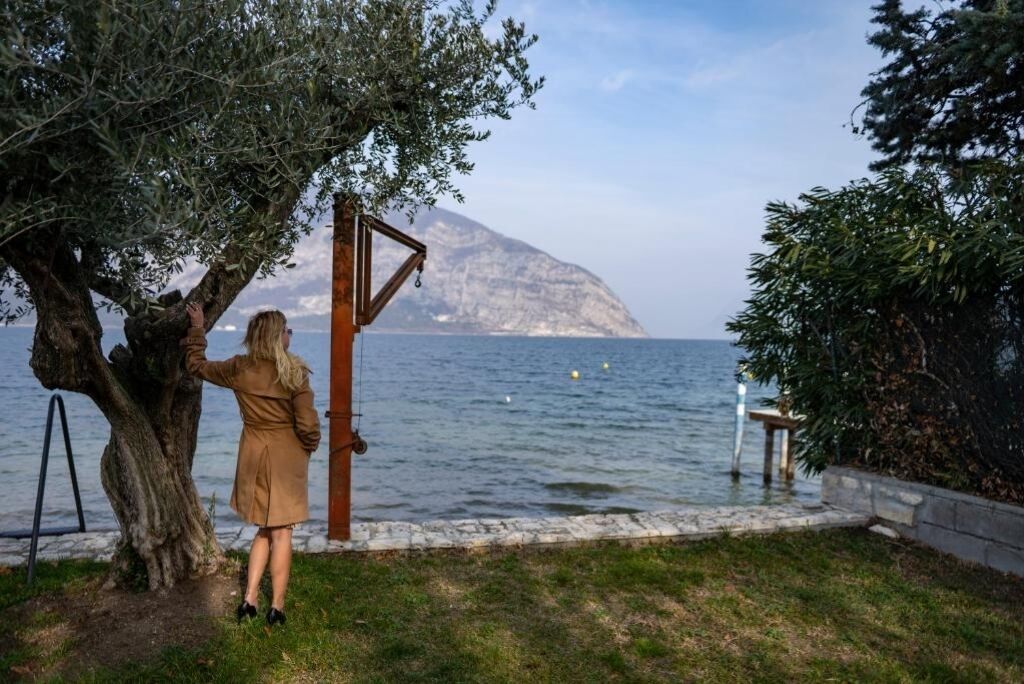 A coastal scene with a person leaning on a tree, a wooden structure, and a view of the sea and a distant mountain.