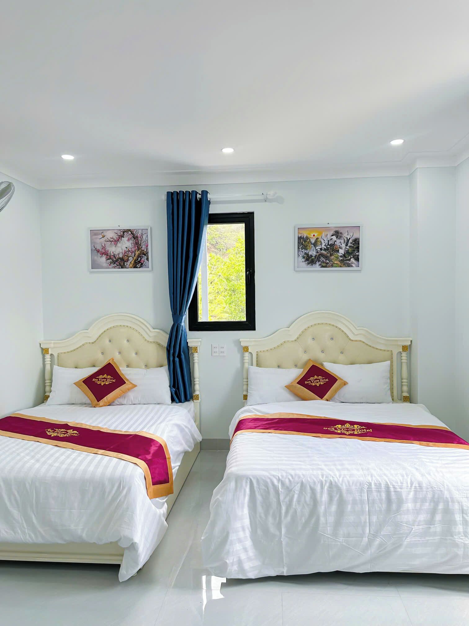 Quarto de hotel com duas camas, cada uma com colcha branca e almofadas bordô, uma janela com cortinas azuis e duas pinturas penduradas na parede.